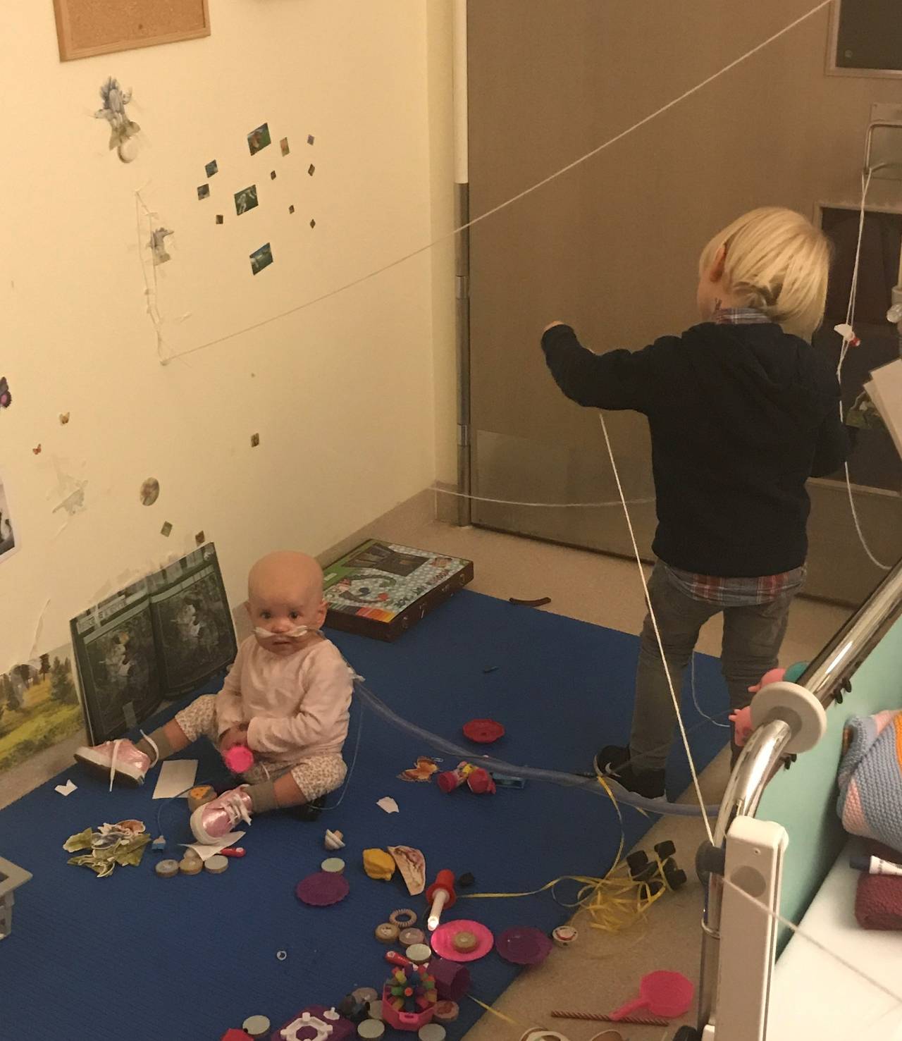 Two children playing on a blue rug on the floor.  A child standing and pulling some strings and one girl sitting on the floor looking at the camera.  She has a tube inserted into her nose.